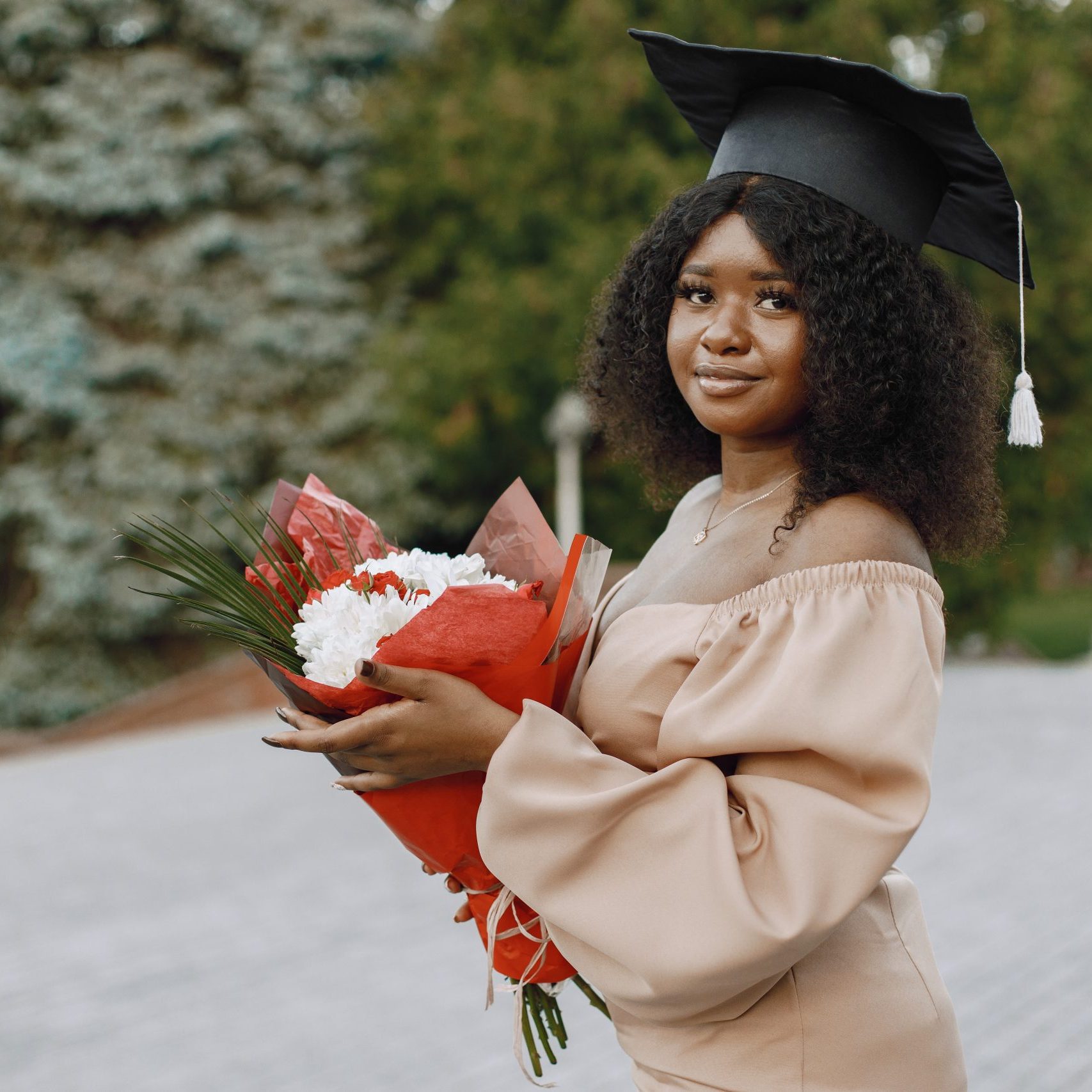 Young afro american female student dressed in beige dress and academic cap. Campus as a background. Girl posing for a photo and holding a flowers.